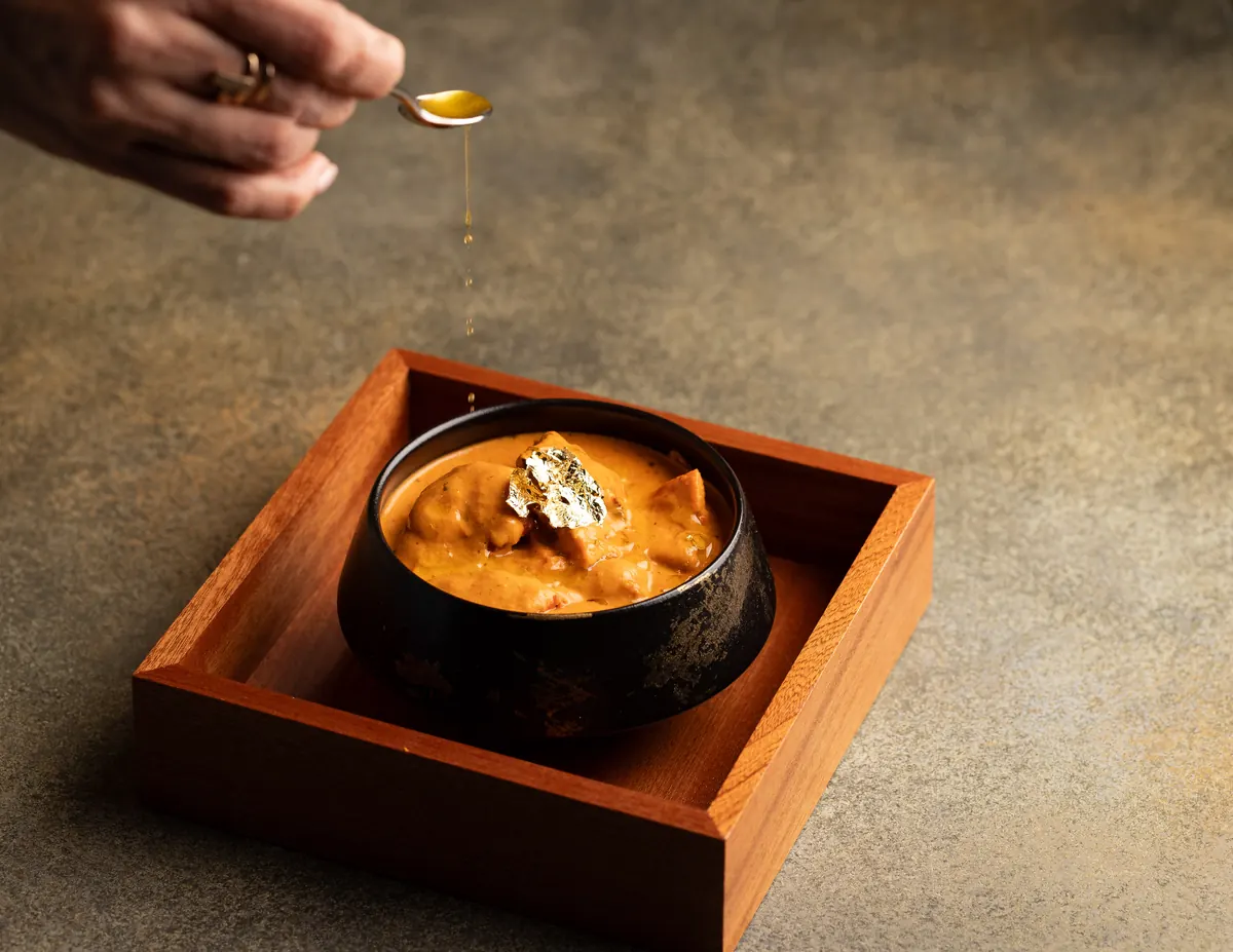 Butter chicken in a black bowl, set in a wooden box, with a spoon of butter poured over it