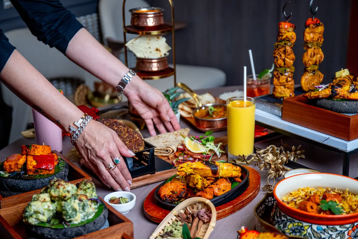 Hands arranging a full table of tandoor dishes, skewers and drinks