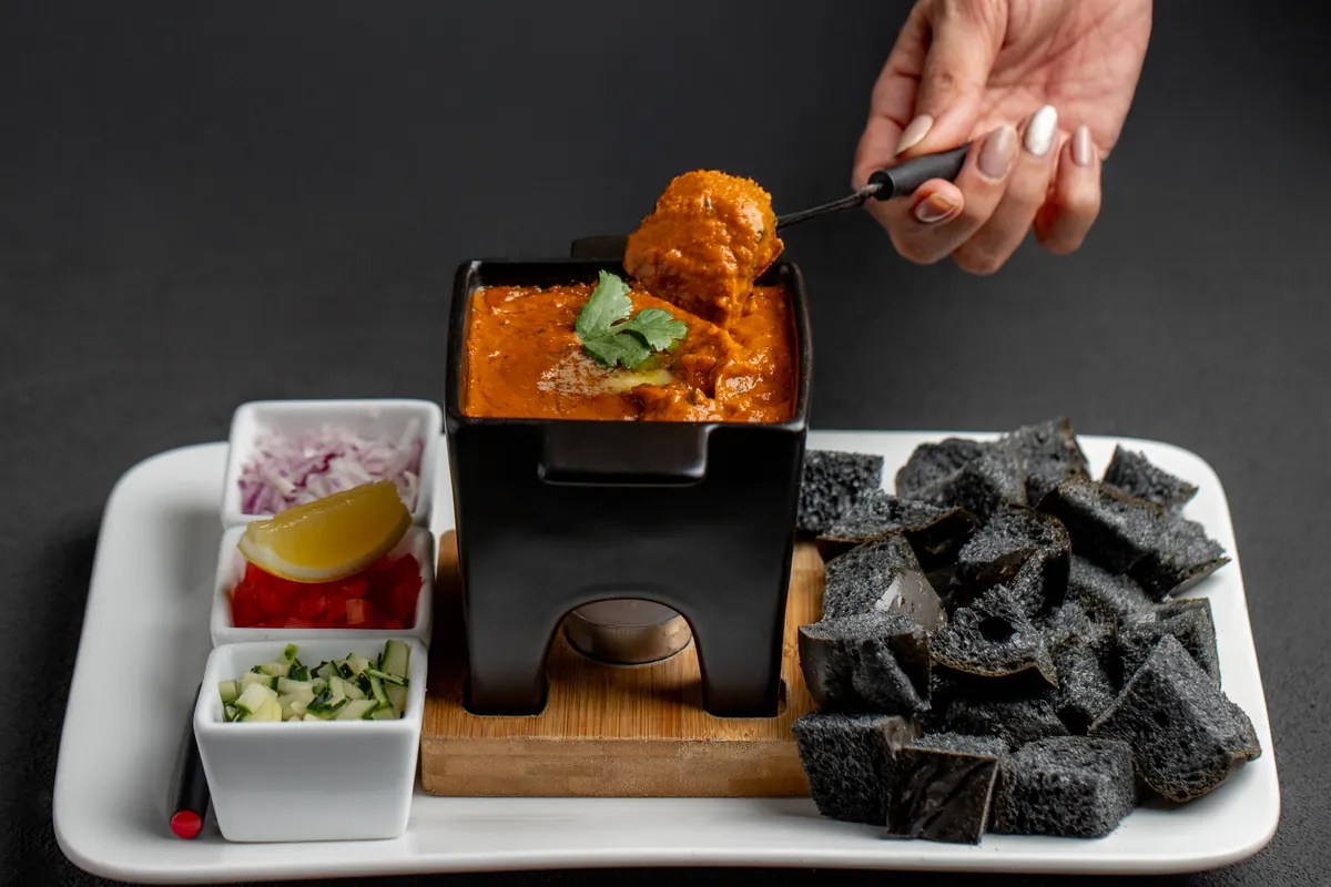Tagline Paav Bhaji Fondue: a hand dipping charcoal pav into a pot of bhaji